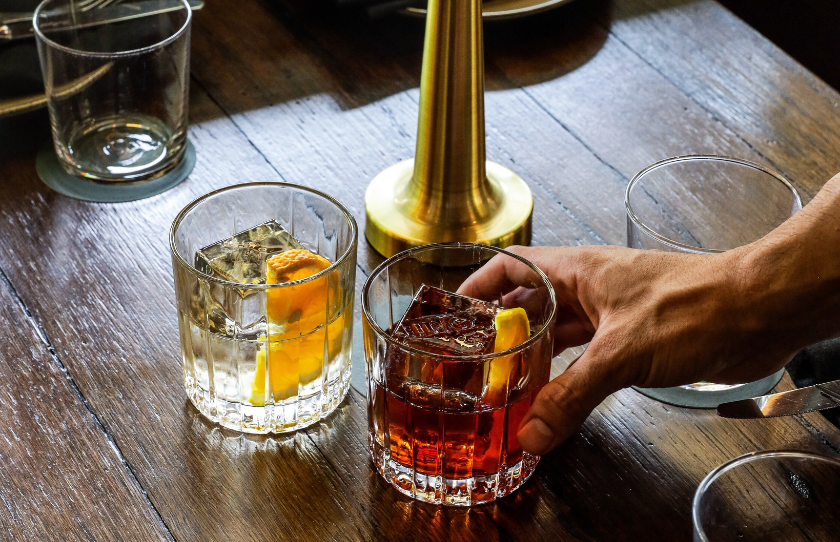 Negroni glasses, one full and one empty. Hand grabbing the filled glass. Gold lamp in the background, all on a wooden table.