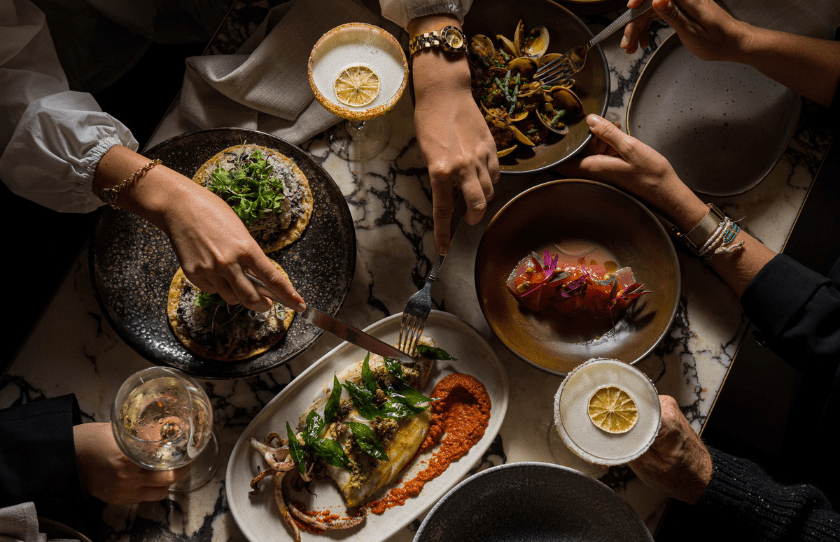 Overshot of Mexican dishes in Esteban with hands cutting into the food and cocktails