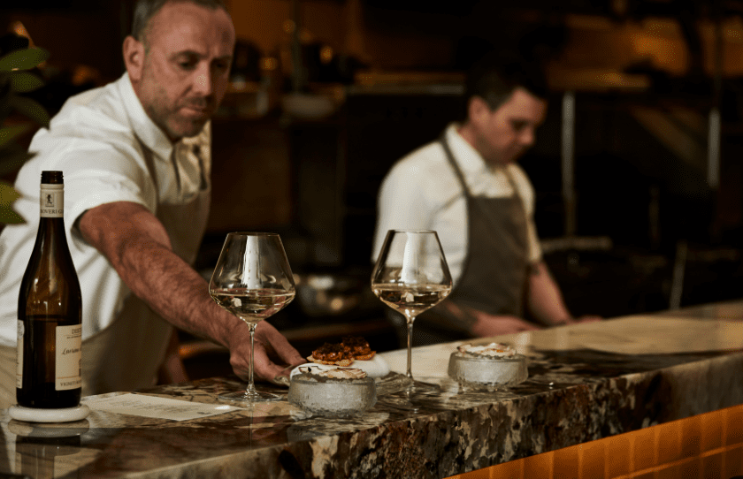 Chefs Joel Bickford and head chef Bradat kitchen pass serving food. bottle of wine with two full glasses on the table at Shellhouse Sydney