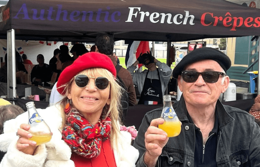 Man and woman at the French Markets holding a drink of orange soda wearing berets and sitting in front of an authentic crepe stand 