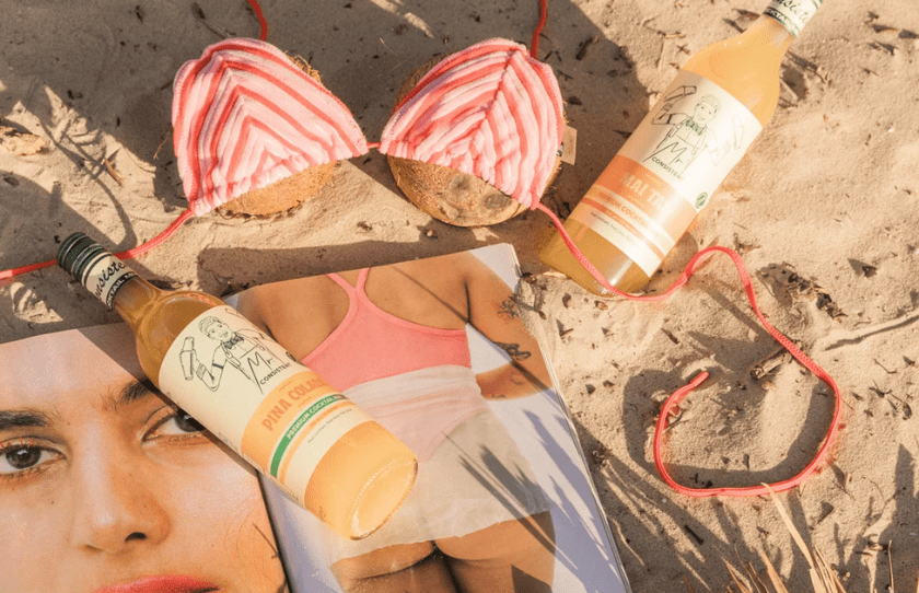 Mr Consistent Pina Colada and Mai Tai from Tiki Collection laid out on the sand with magazines, a cocnut and bikini top for October 2024 Drinks guide