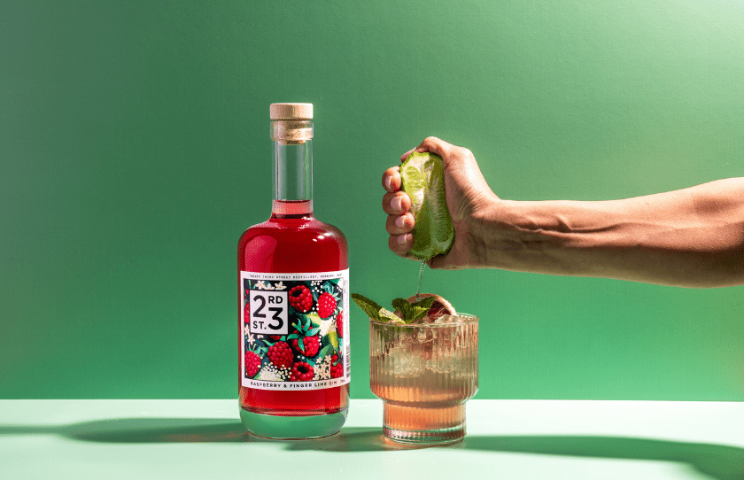 23rd Street Distillery New Raspberry & Finger Lime Gin with a glass next to it and a lime being squeesed in. October 2024 Drinks Guide