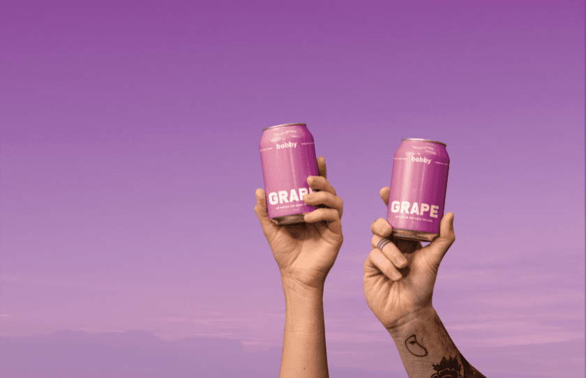 Two hands holding up a grabe bobby can each with a purple backdrop - October 2024 Drinks Guide